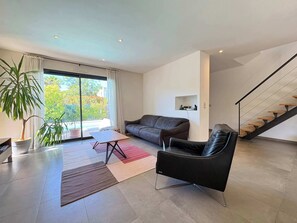 A modern living room with a sofa, a coffee table, a chair, a staircase, and a large window with a view of greenery. - Villa Salina (Agde)