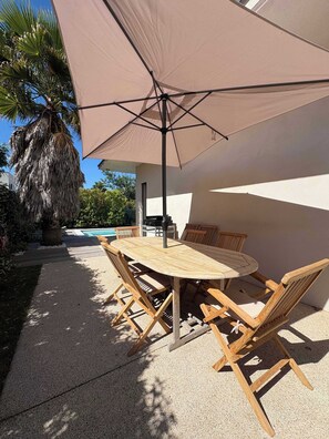 An outdoor patio with a round wooden table and chairs, a large beige umbrella, and a pool in the background. - Villa Salina (Agde)