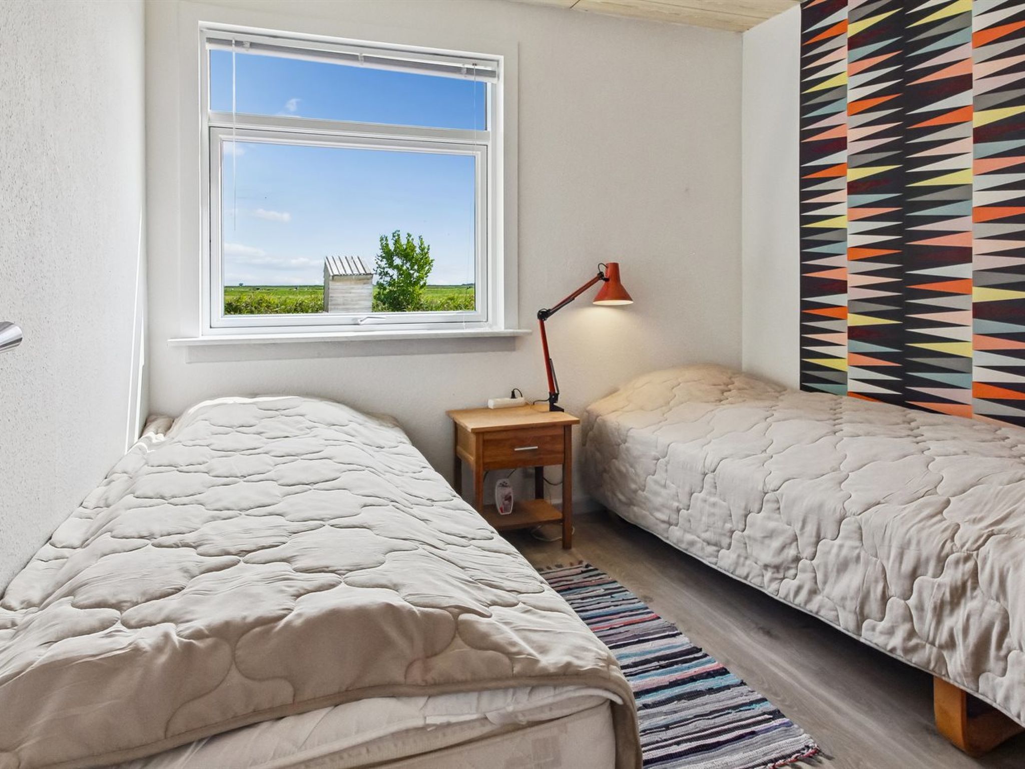 Une chambre avec deux lits, une fenêtre donnant sur un paysage verdoyant, une table de chevet avec une lampe et un mur décoré d’un motif.