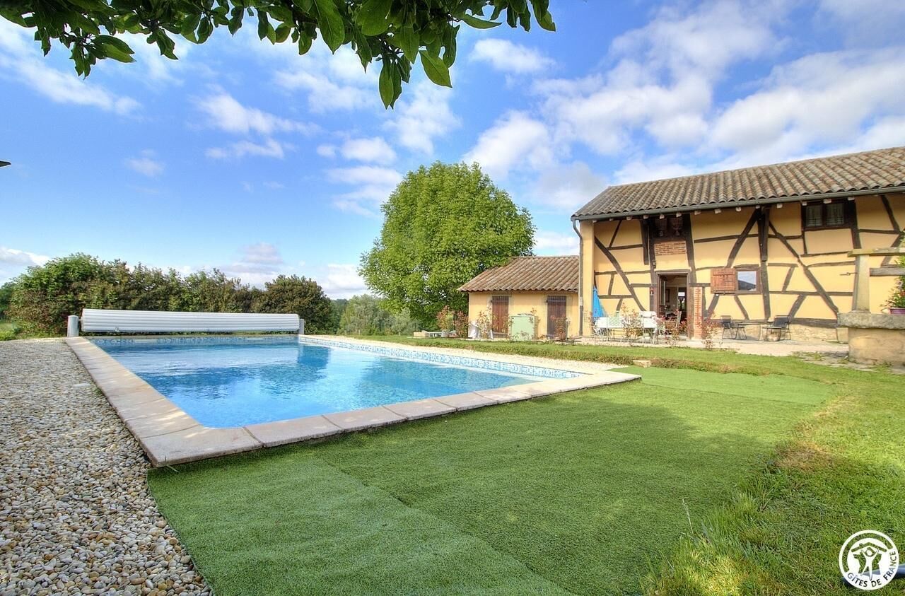 Piscina com área gramada ao redor, um edifício tradicional com telhado em azulejos e uma grande árvore ao fundo.