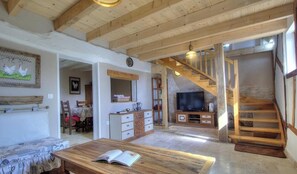 Un intérieur rustique avec des poutres apparentes, un coin repas, un téléviseur et un escalier.