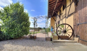 Un bâtiment doté d’une grande roue en bois, d’une pergola couverte de plantes et d’une cour intérieure recouverte de gravier.