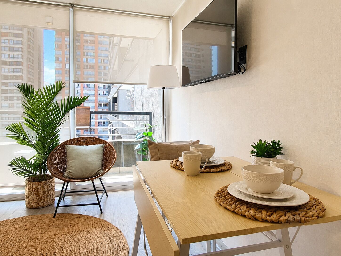 Uma sala de estar moderna com uma janela ampla, uma cadeira de vime, uma mesa de madeira com pratos decorativos e uma televisão na parede.