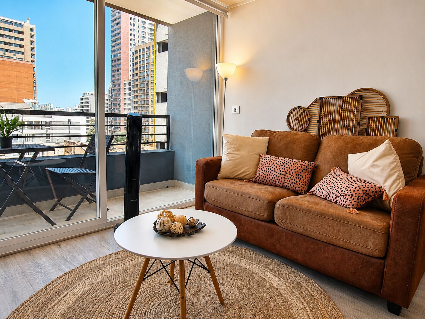 A modern living room with a brown sofa, a white coffee table, and a balcony with a view of tall buildings.