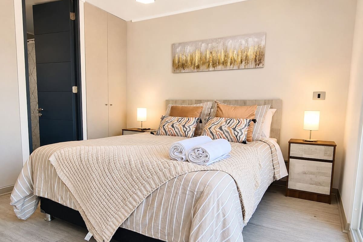 A neatly made bed with striped bedding, two bedside tables with lamps, a door, and a wall-mounted abstract art piece.