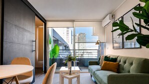 Una sala de estar moderna con un sofá, una mesa de comedor, sillas, una mesita con una planta y un balcón con vistas a edificios.