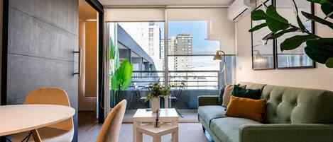 A modern living room with a sofa, a dining table, chairs, a small table with a plant, and a balcony with a view of buildings.