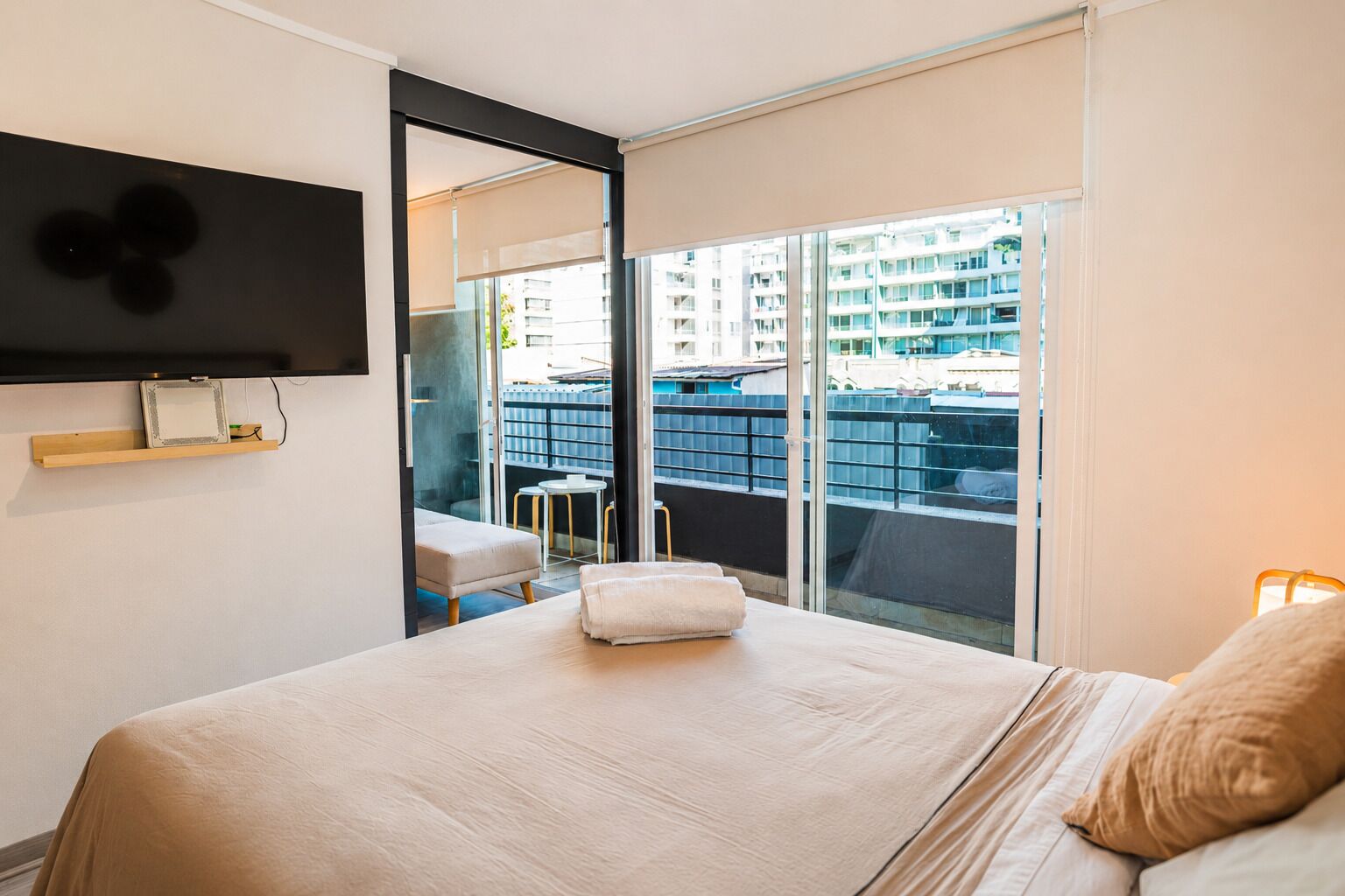 A hotel room with a bed, a TV mounted on the wall, a balcony with a view of buildings, and a small table with chairs.