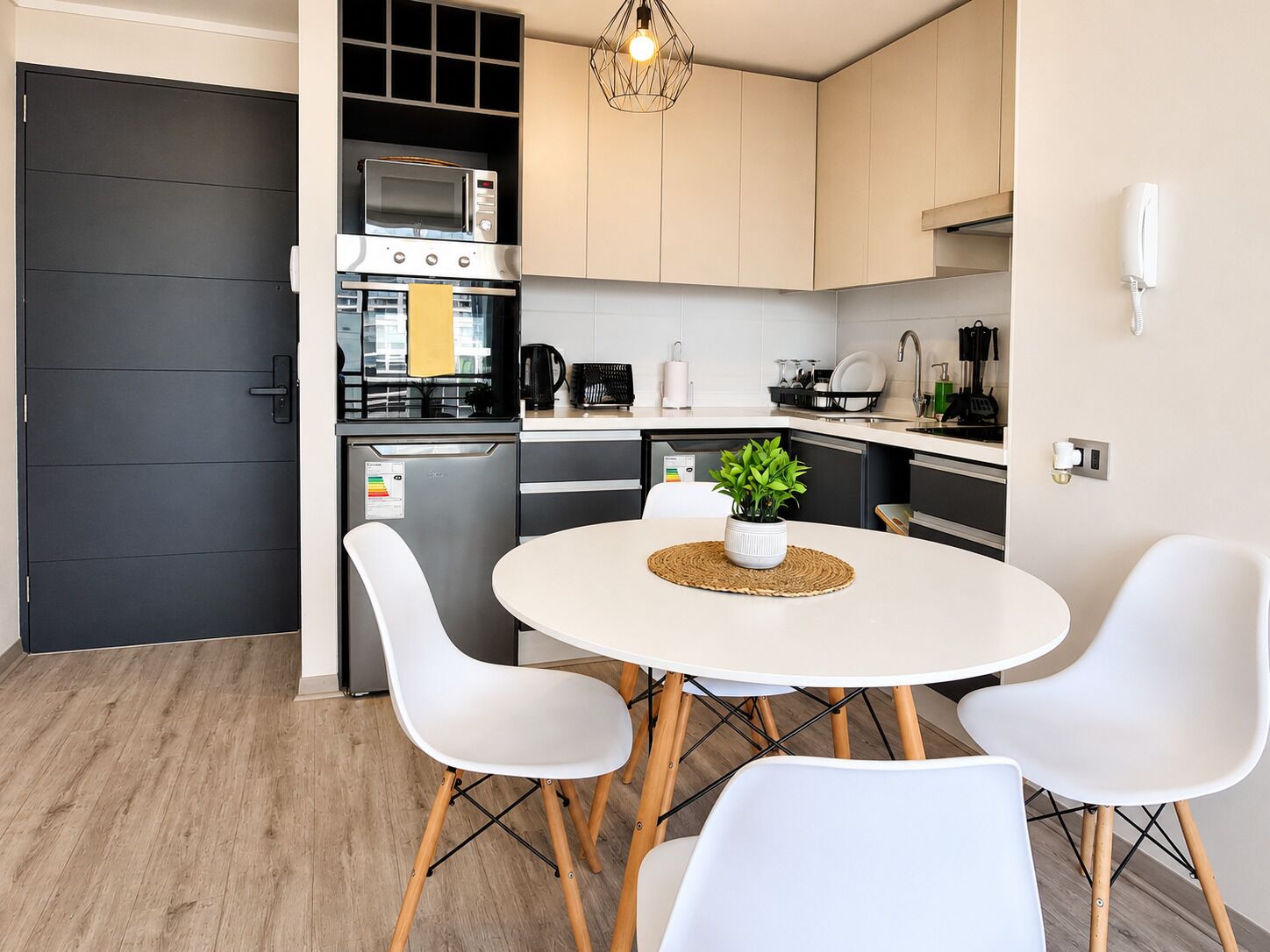 A modern kitchen with a built-in oven, microwave, and coffee maker, a dining area with a round table and white chairs, and a small potted plant on the table.