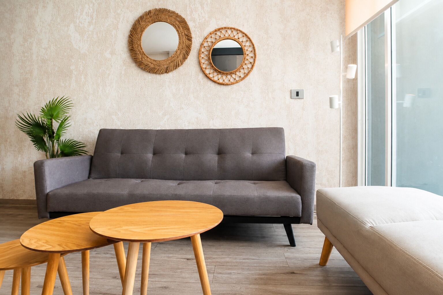 A modern living room with a grey sofa, two round wooden tables, and two wall-mounted mirrors.