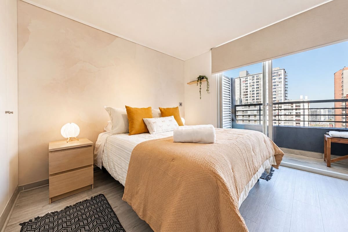 A bedroom with a bed, bedside table, lamp, and a view of buildings through a large window.