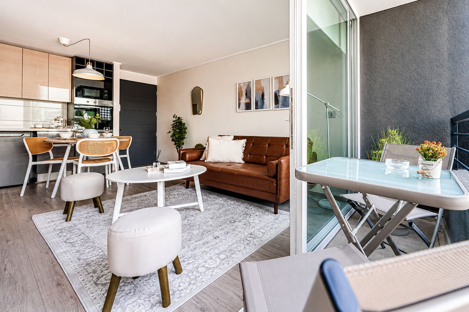 Una sala moderna con sofá, mesa de centro y sillas, una cocina con armarios y electrodomésticos, y un balcón con una mesa y planta en maceta.