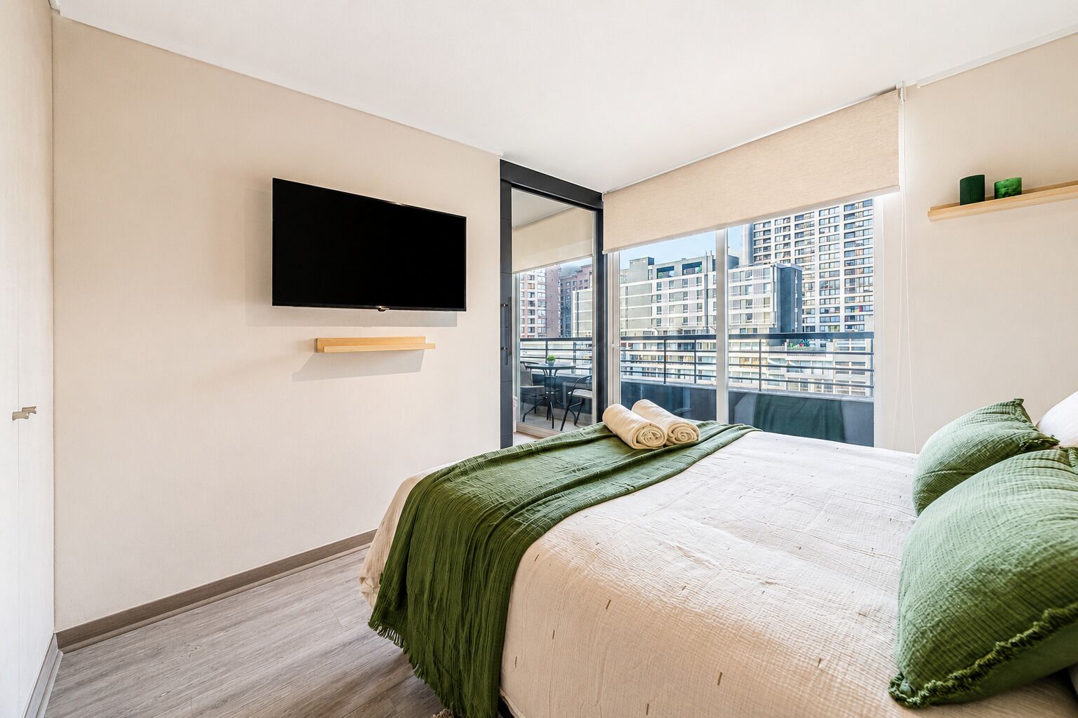 A bedroom with a bed, a TV mounted on the wall, a window with a city view, and a small shelf with items.