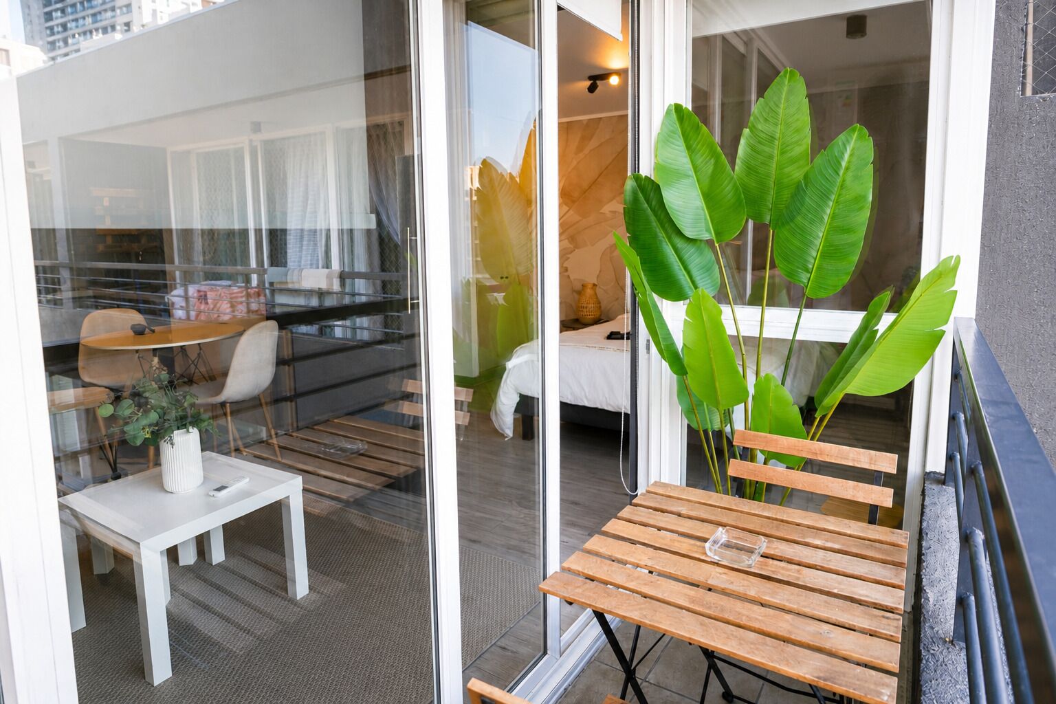Moderni hotellihuone, jossa on lasiovi, joka johtaa parvekkeelle. Huoneessa on puinen pöytä, valkoinen tuoli ja suuri viherkasvi.