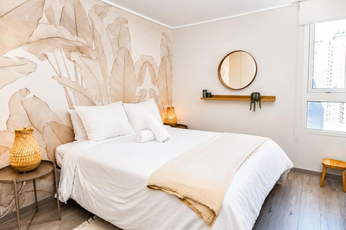 A bedroom with a large bed, a round mirror, a shelf with plants, and a window.
