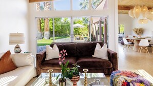 Ein großzügiger Wohnbereich mit einem braunen Sofa, einem Glastisch und Blick auf einen Garten durch große Fenster.