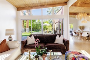 A spacious living area with a brown sofa, a glass coffee table, and a view of a garden through large windows. - The Retreat, Private MidCentury home in sunny Palm Springs (Palm Springs)