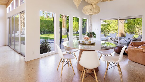 Ein modernes Interieur mit einem runden Esstisch, weißen Stühlen, einer gewebten Deckenleuchte und großen Fenstern mit Blick auf einen Garten.