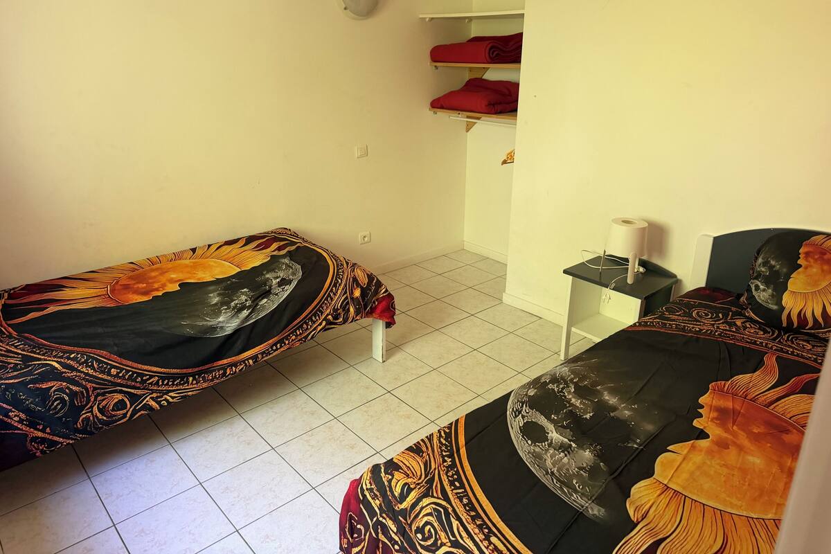 A room with two beds, a small table, and a shelf with red pillows.