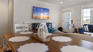 Un área de comedor con una mesa de madera, individuales blancos, un jarrón de cristal decorativo y un cuadro de un atardecer en la pared.