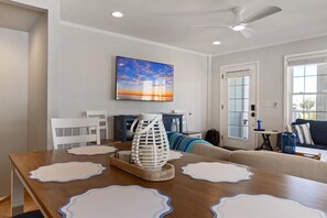 A dining area with a wooden table, white placemats, a decorative glass vase, and a painting of a sunset on the wall. - Steps to beach | Ocean Views | Beach Gear (Carolina Beach)