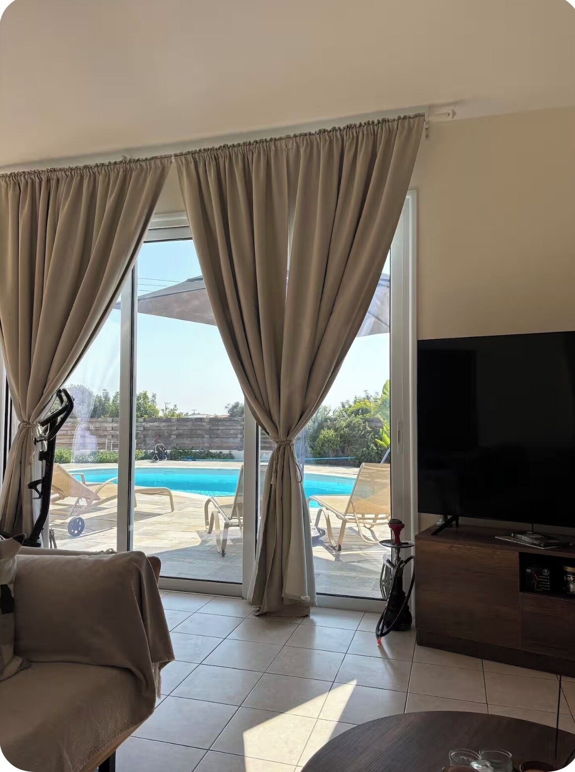 Une chambre dotée d’une télévision, d’un canapé et offrant une vue sur la piscine grâce à de grandes fenêtres équipées de rideaux.