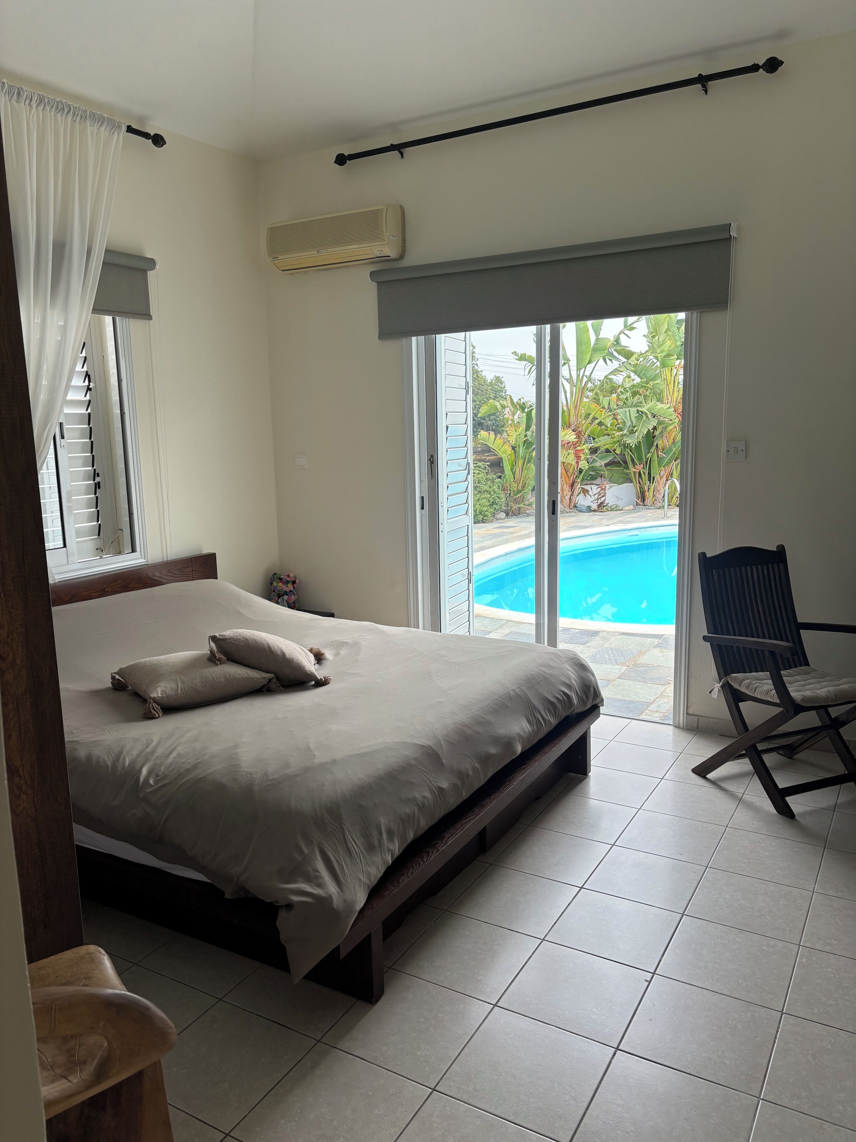 A bedroom with a bed, a chair, a window with curtains, an air conditioning unit, and a view of a swimming pool.