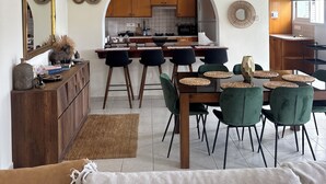 A spacious living area with a dining table, green chairs, and a sofa. A kitchen with wooden cabinets and a hanging light fixture is visible in the background. - RELAX IN A PRIVATE 3 BEDROOMS BUNGALOW WITH SWIMMING POOL. (Paphos)