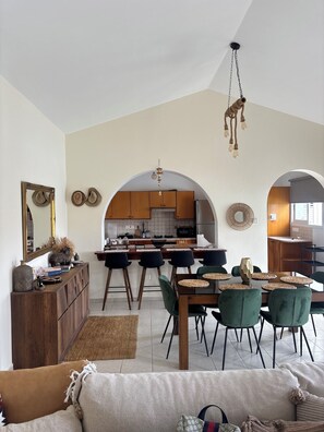 A spacious living area with a dining table, green chairs, and a sofa. A kitchen with wooden cabinets and a hanging light fixture is visible in the background. - RELAX IN A PRIVATE 3 BEDROOMS BUNGALOW WITH SWIMMING POOL. (Paphos)