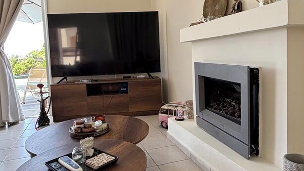A living room with a flat-screen TV, a fireplace, a round table with a tray, and a clock on the wall. - RELAX IN A PRIVATE 3 BEDROOMS BUNGALOW WITH SWIMMING POOL. (Paphos)