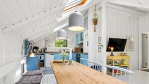 Un vaste espace dédié à la salle à manger, avec une table en bois, des bancs rembourrés, un éclairage suspendu et, au fond, une vue sur la cuisine.