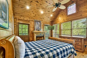 Ein Schlafzimmer in einer Holzhütte mit Bett, Kamin, Deckenventilator und großen Fenstern.