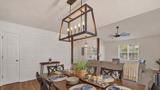 A dining area with a wooden table set for six, a hanging light fixture, and a view to a living room with a sofa and a window. - Lake Life Chapin Retreat, Sleeps 7, King Bed (Chapin)
