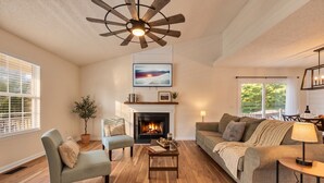 A living room with a fireplace, a ceiling fan, a sofa, a coffee table, and a TV mounted above the fireplace. - Lake Life Chapin Retreat, Sleeps 7, King Bed (Chapin)