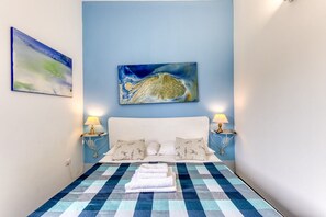 Ein Bett mit kariertem Überwurf, zwei Nachttischlampen, ein weißes Kopfteil und zwei an der Wand befestigte Bilder.