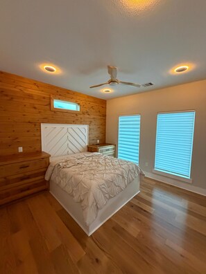 Ein Schlafzimmer mit einem Bett, einem Holz-Kopfende, Nachttischen, einem Deckenventilator und zwei Fenstern mit Jalousien.
