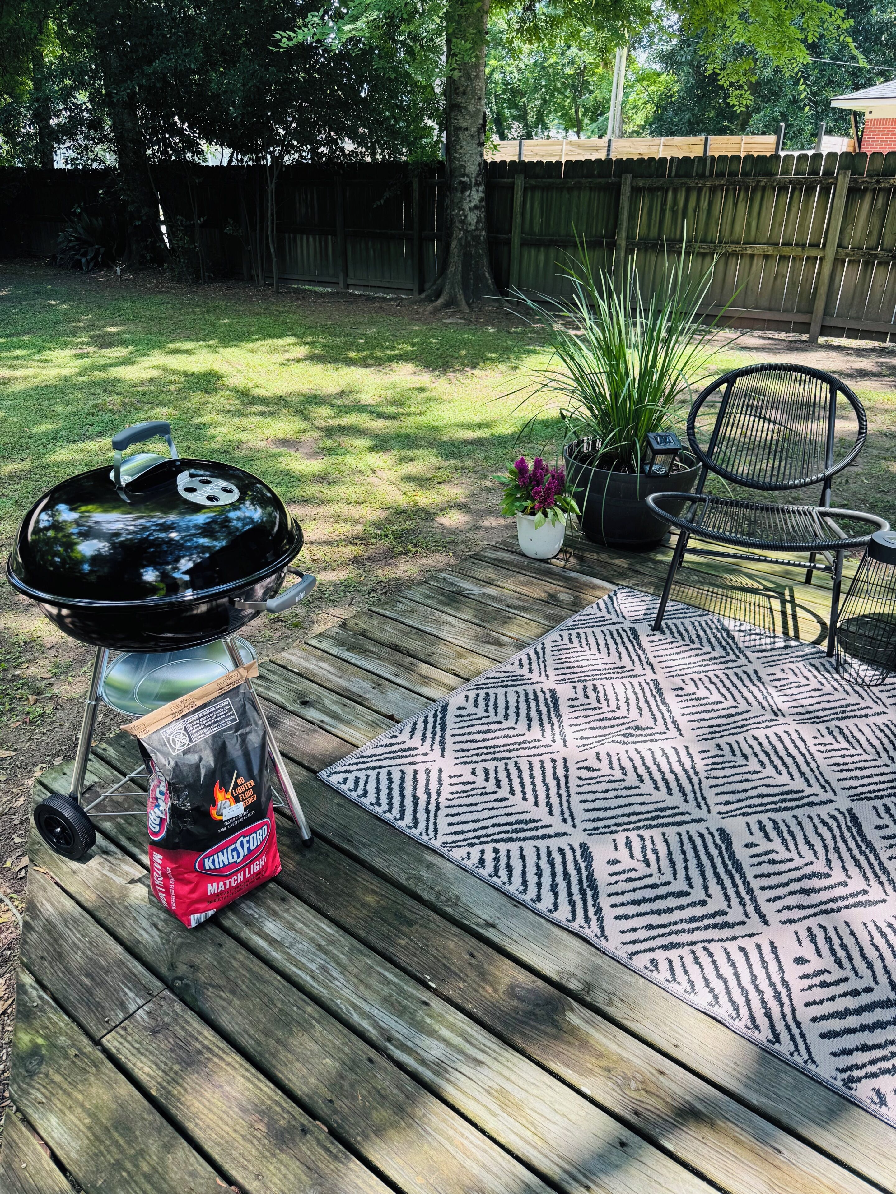 Un patio trasero con una parrilla, una silla, una planta en maceta y un tapete estampado.