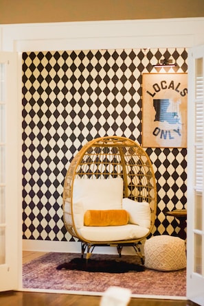 Una habitación con papel tapiz estampado, una silla de mimbre con cojines, una alfombra pequeña y un cartel enmarcado en la pared.