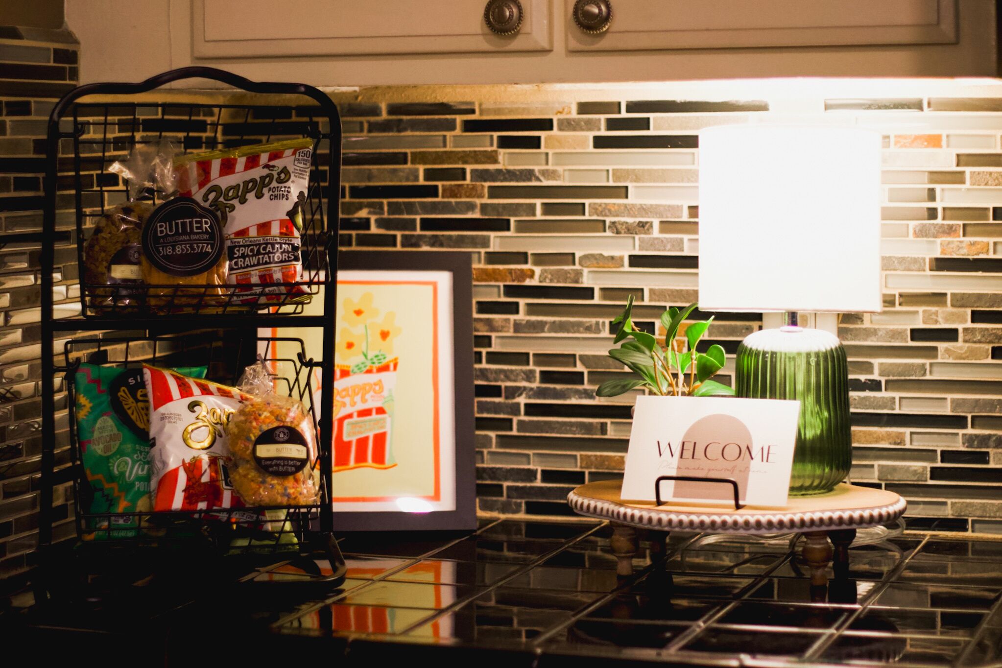 Una encimera de cocina con un estante metálico que contiene aperitivos, un cuadro enmarcado y un cartel de bienvenida sobre una pequeña mesa.