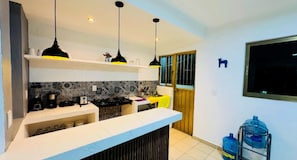 Una cocina moderna con encimera blanca, lámparas colgantes negras y amarillas, salpicadero de azulejos y ventana con vistas.