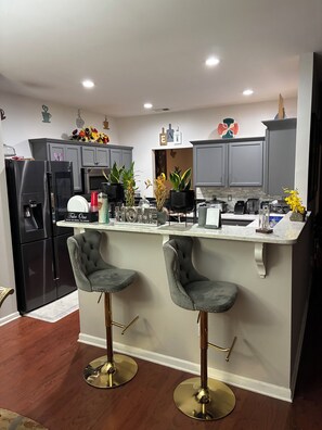 A modern kitchen with grey cabinets, a black refrigerator, two bar stools, and a central island with a sink and countertop. - Stunning 5-BedroomHome with Space,Style & Comfort (Fairburn)
