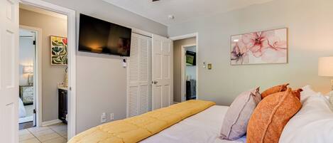 Un dormitorio con cama, ventilador de techo, televisor montado en la pared y puerta que da a otra habitación.