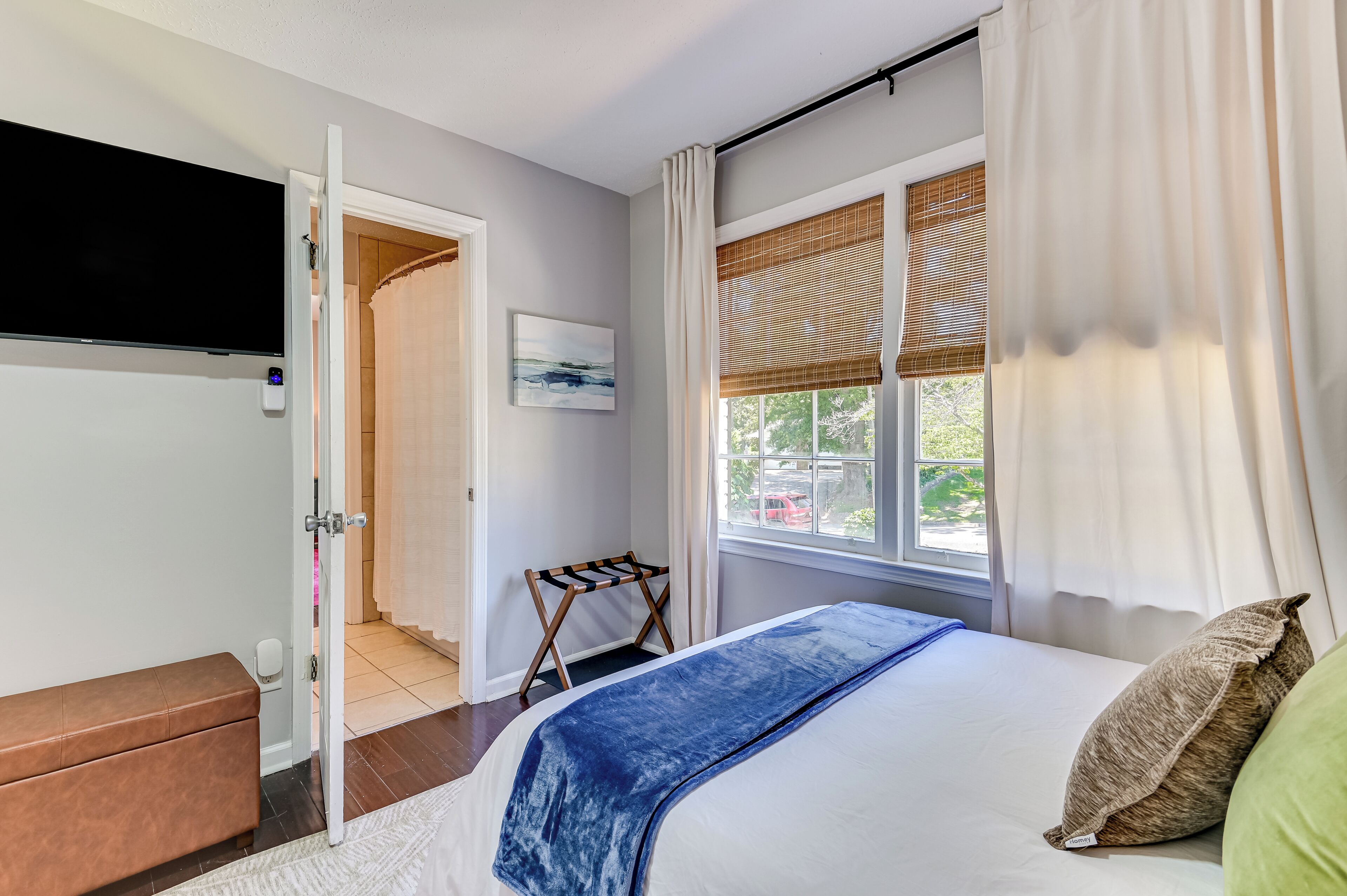 A bedroom with a bed, a bench, a TV, a window with curtains, and a door leading to another room.