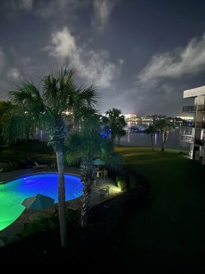 A nighttime view of a swimming pool area with palm trees and a view of a cityscape in the background. - Bayfront Condo with Peaceful Water Views (Fort Walton Beach)