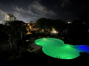 Vista notturna di una piscina illuminata di verde, circondata da alberi e con un edificio sullo sfondo.