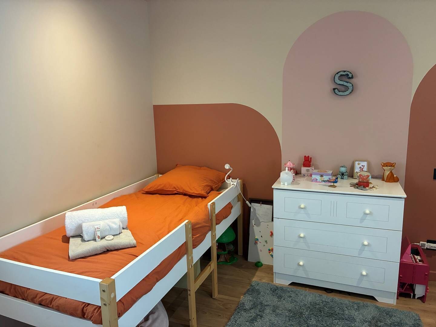 A single-bed room with a white dresser, a small desk, and a wall-mounted letter 'S'.