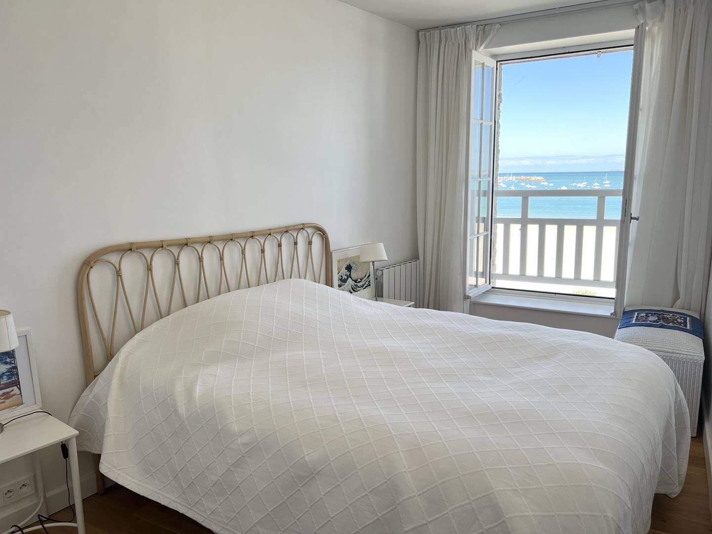 A bedroom with a bed, a bedside table, a lamp, a window with curtains, and a view of the sea.