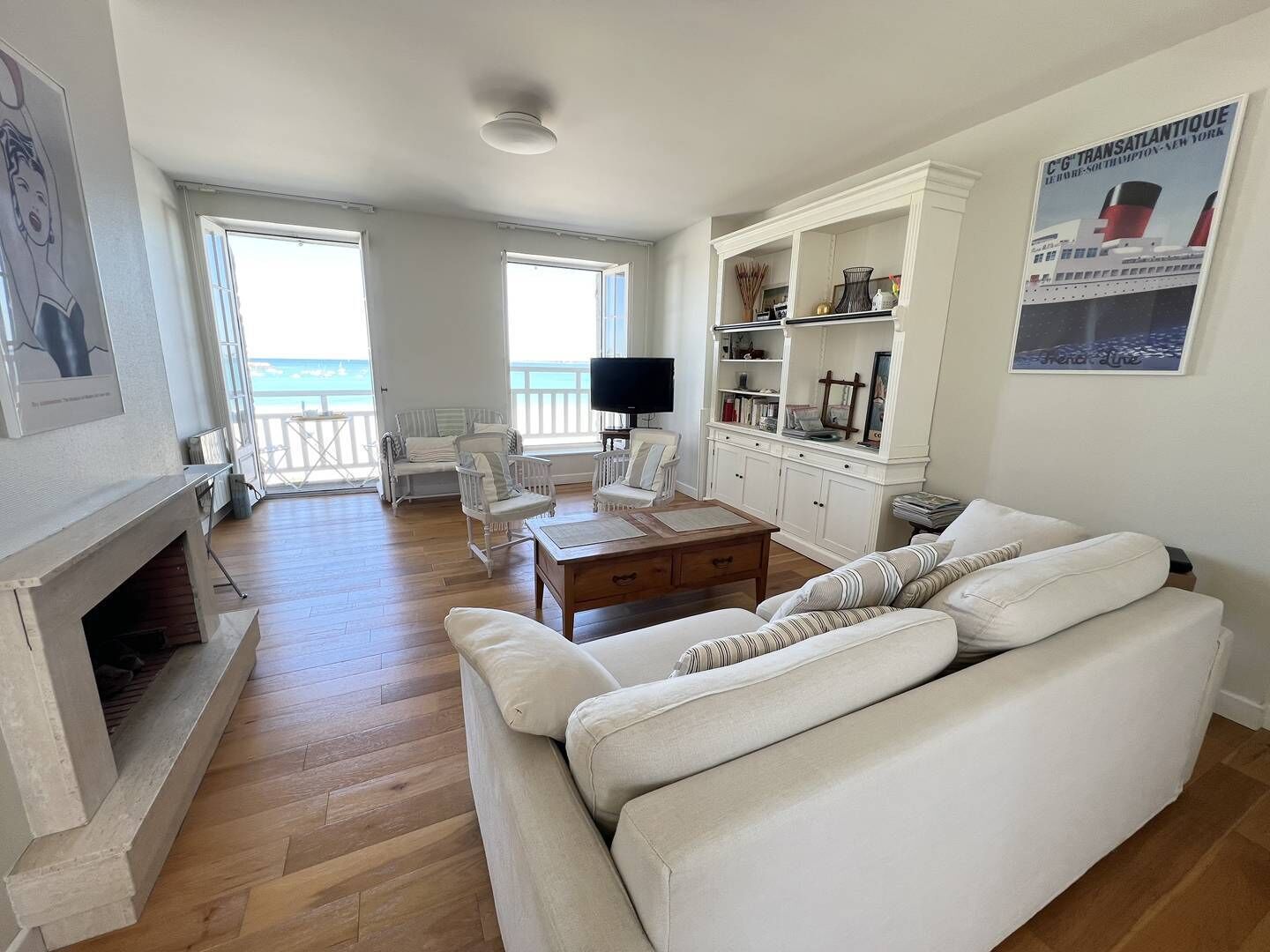 Una sala de estar con chimenea, sofás blancos, una mesa de centro y un televisor. Hay dos ventanas con vista al mar.