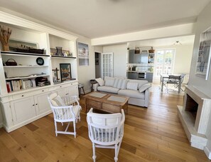 Ein geräumiges Wohnzimmer mit einer Couch, zwei weißen Sesseln, einem Couchtisch und einem Kamin.