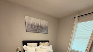 Un dormitorio con cama, una mesita de noche, lámpara, ventana con persianas y un cuadro en la pared.